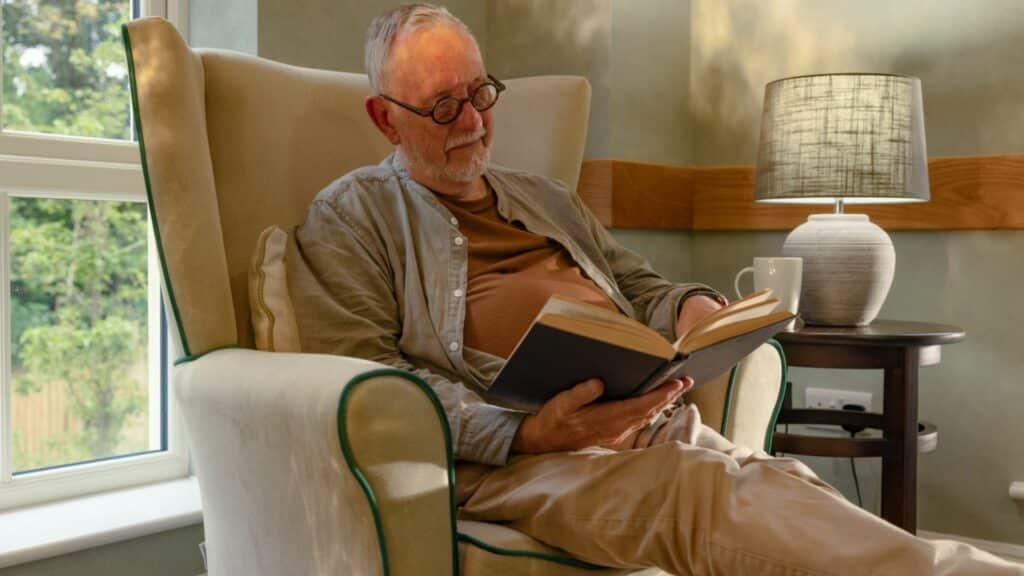 Elderly man reading book by window lamp.